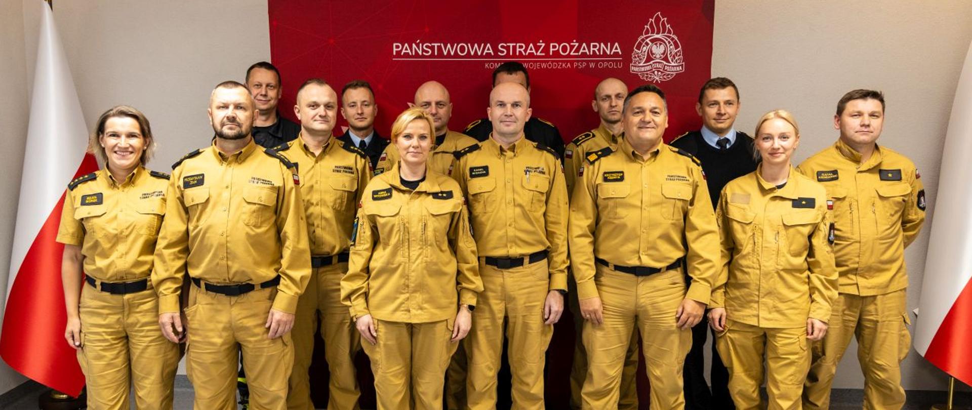Wspólne zdjęcie członków komisji dyscyplinarnej z Opolskim Komendantem Wojewódzkim PSP. Strażacy w ubraniach służbowych stoją na tle banneru Państwowej Straży Pożarnej (koloru czerwonego) oraz dwóch Flag RP.