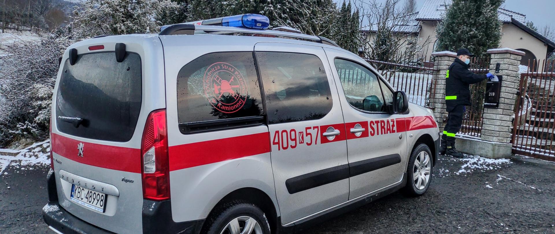 Mężczyzna ubrany w czarny mundur strażacki z odblaskowymi paskami, czarna czapką na głowie, czarnym butami, na rękach ma założone rękawiczki lateksowe koloru niebieskiego, na twarzy ma ubrana maseczkę chirurgiczną koloru biało niebieskiego , stoi on na asfalcie , ręce ma położone na skrzynce pocztowej koloru czarnego i wrzuca do niej ulotkę informacyjną. Ogrodzenie jest koloru brązowego a murki ogrodzeniowe koloru białego. Na nimi znajduje się budynek mieszkalny koloru białego, dach budynku pokryty jest śniegiem. Za mężczyzną stoi pojazd pożarniczy koloru srebrnego z czerwonym paskiem. Na drzwiach bocznych znajduje się napis Straż oraz numery operacyjne 409 k 57 na dachu pojazdu belka świateł błyskowych. Z tył widoczny napis Peugeot oraz rejestracja z numerami KBC 48998. Za pojazdem krzewy na których znajduje się śnieg.