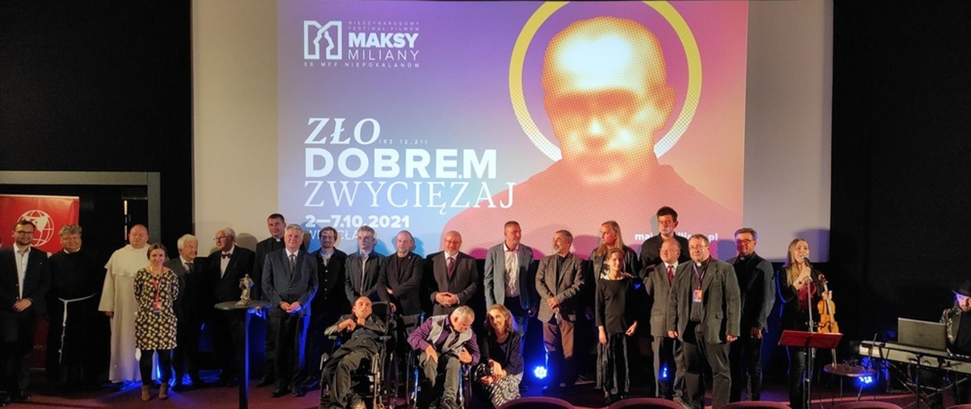 Zdjęcie w poziomie. Uczestnicy gali stojący na tle telebimu z wizerunkiem św. Maksymiliana Kolbe