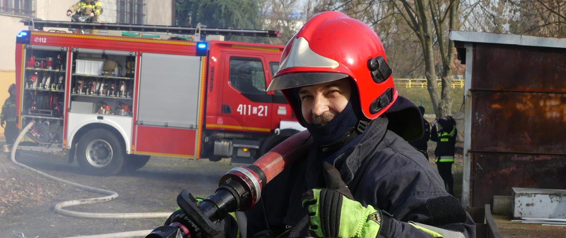 Zdjęcie przedstawia asp. sztab. Roberta Maciejewskiego ubranego w ubranie specjalne i hełm strażacki. W rękach trzyma prądownicę. W tle samochód gaśniczy JRG Brodnica.