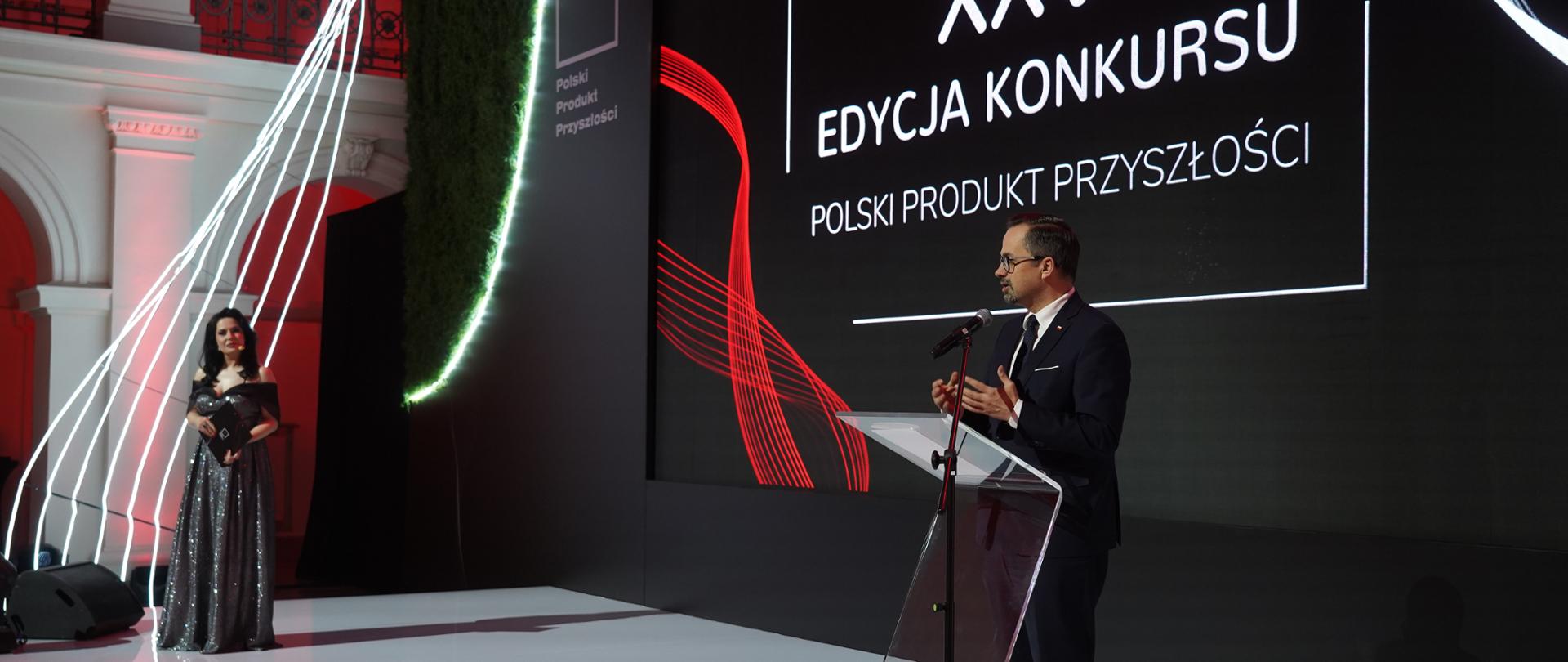Wiceminister Marcin Horała stoi na scenie z prawej strony i mówi do mikrofonu.