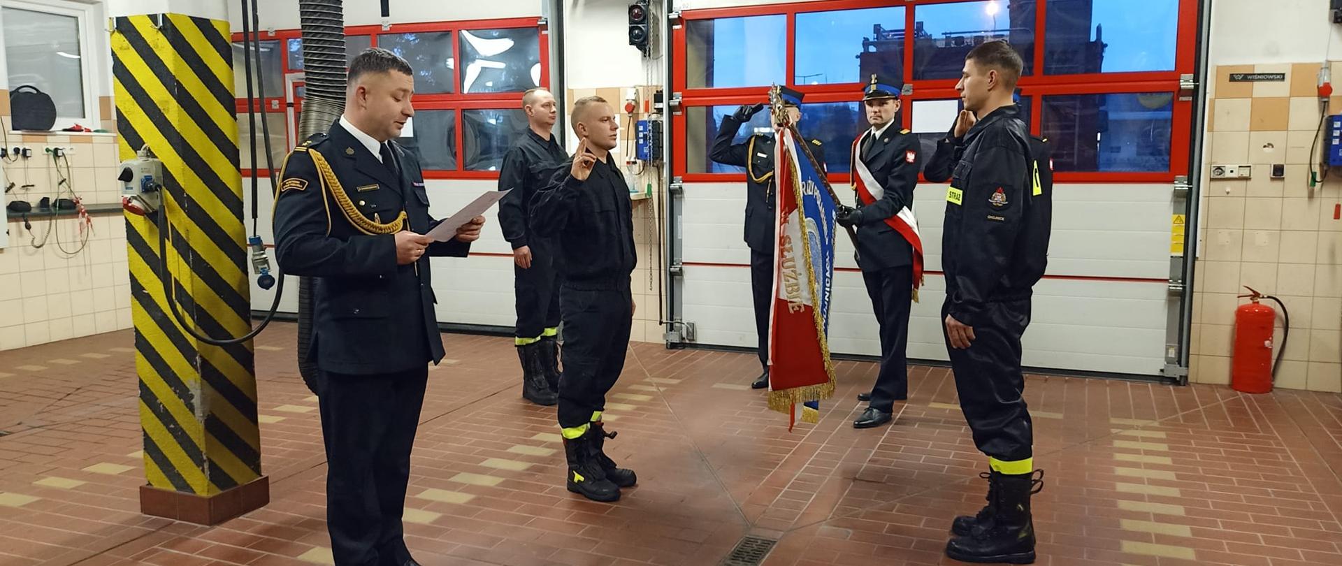 Komendant odczytuje treść roty przysięgi. Strażacy salutują. W tyle, na tle bram garażowych stoi poczet sztandarowy.