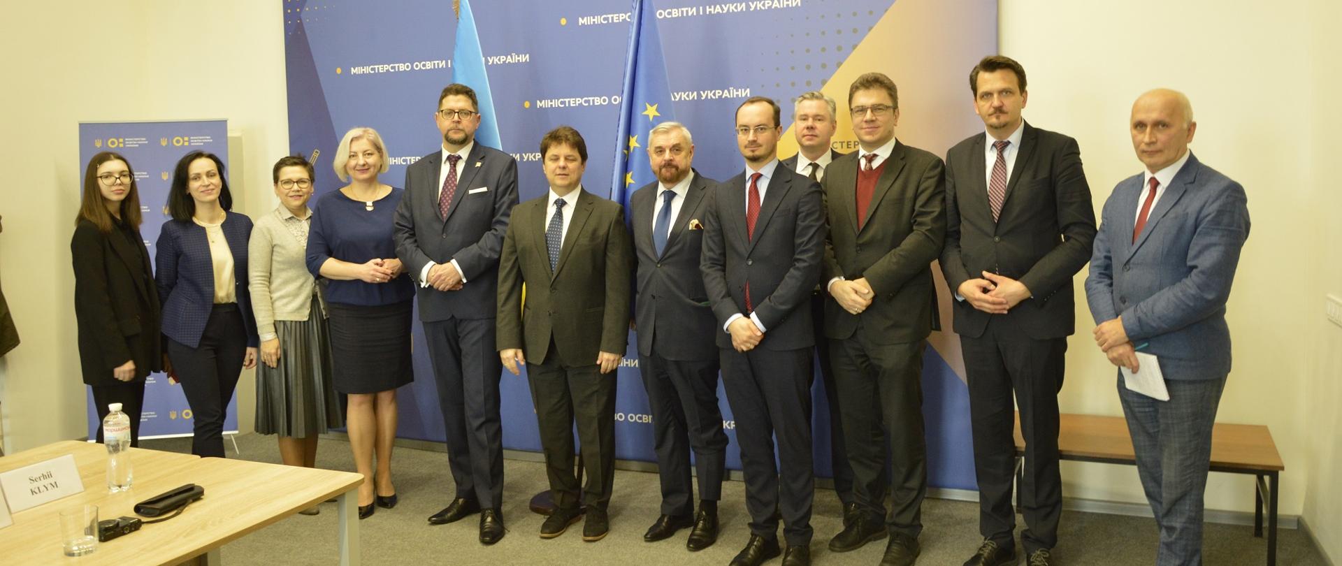 Pod niebieską ścianką z ukraińskimi napisami stoi w rzędzie grupa osób.