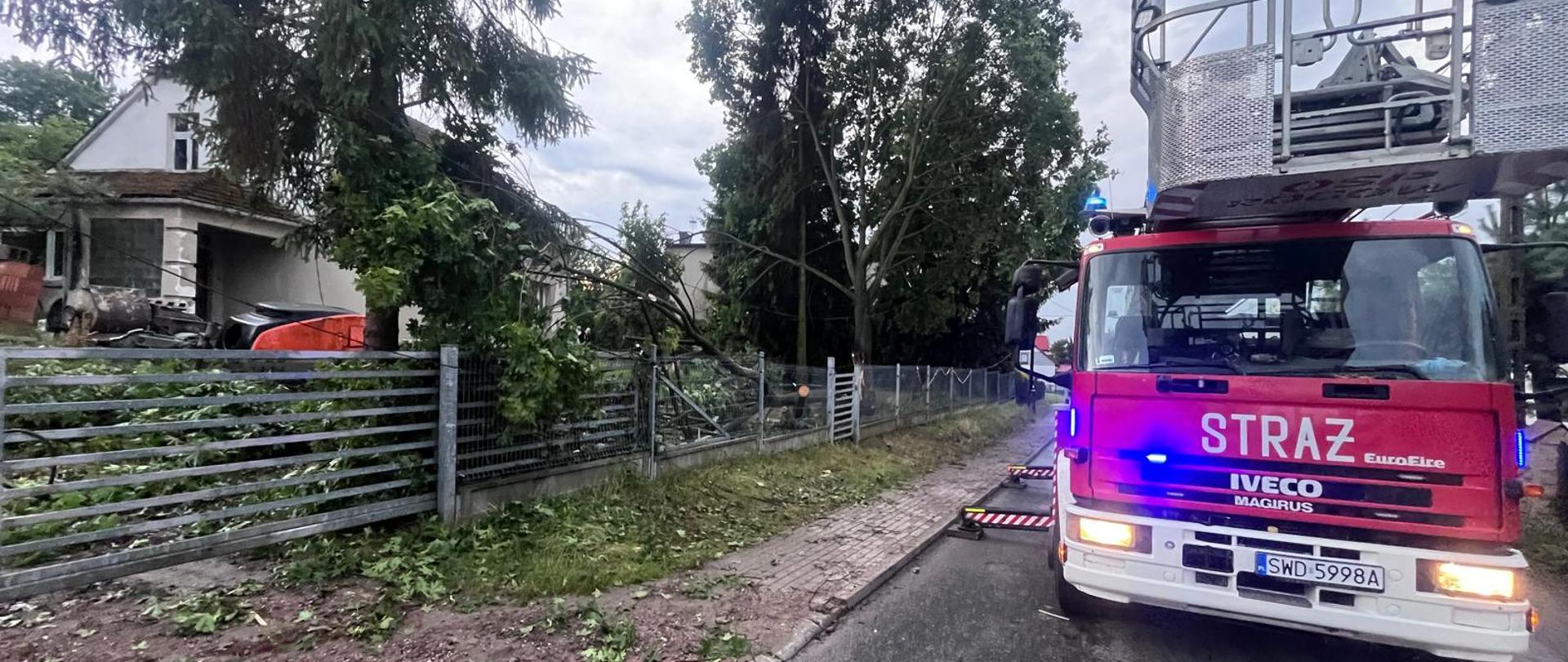 Czerwony samochód strażacki Iveco Magirus z włączonymi sygnałami świetlnymi stoi na drodze podczas akcji usuwania powalonego drzewa, które spadło na ogrodzenie posesji