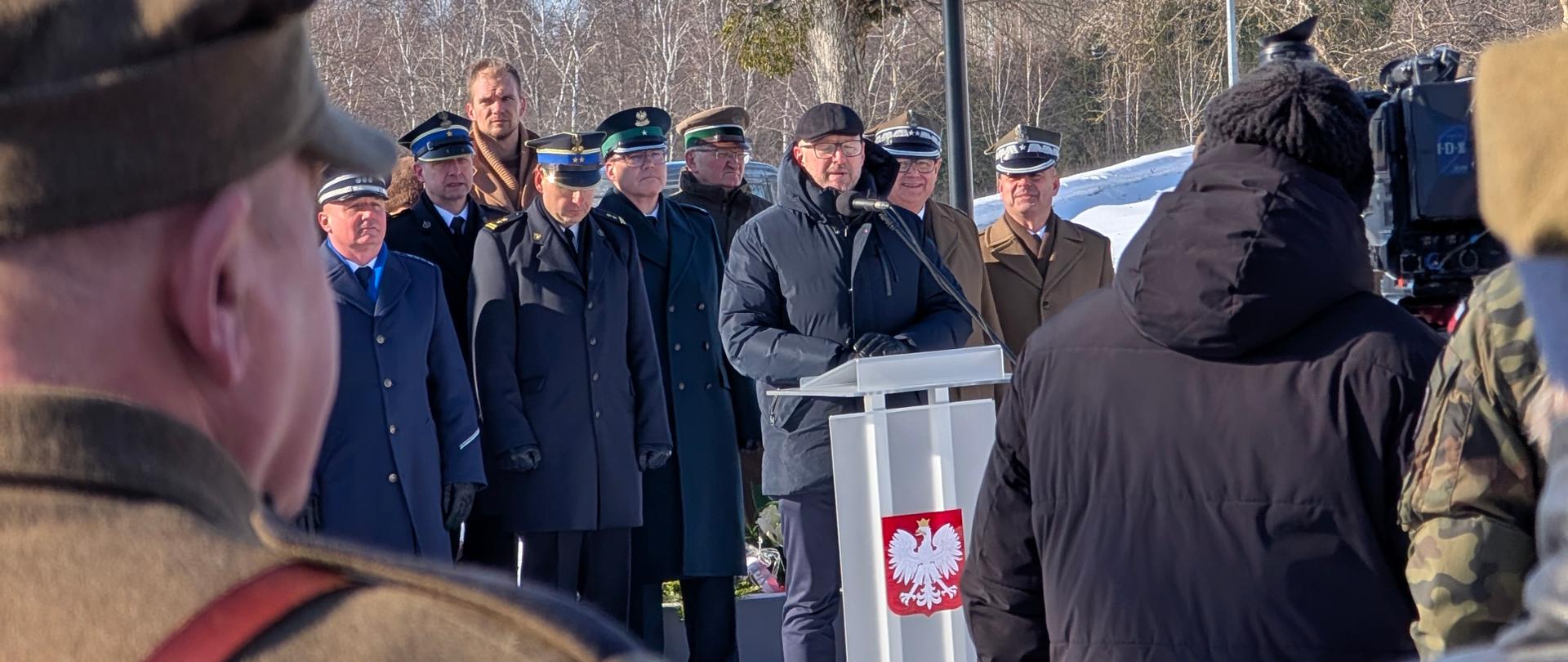 Mężczyzna stoi przy mównicy, grupa osób w mundurach stoi za nim