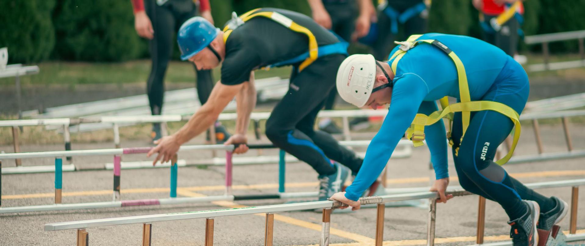 Na zdjęciu widocznych jest dwóch zawodników przygotowujących się do startu w konkurencji wspinanie przy użyciu drabiny hakowej na 3 piętro wspinalni. Zawodnicy znajdują się w blokach startowych, trzymają w dłoniach drabiny hakowe.