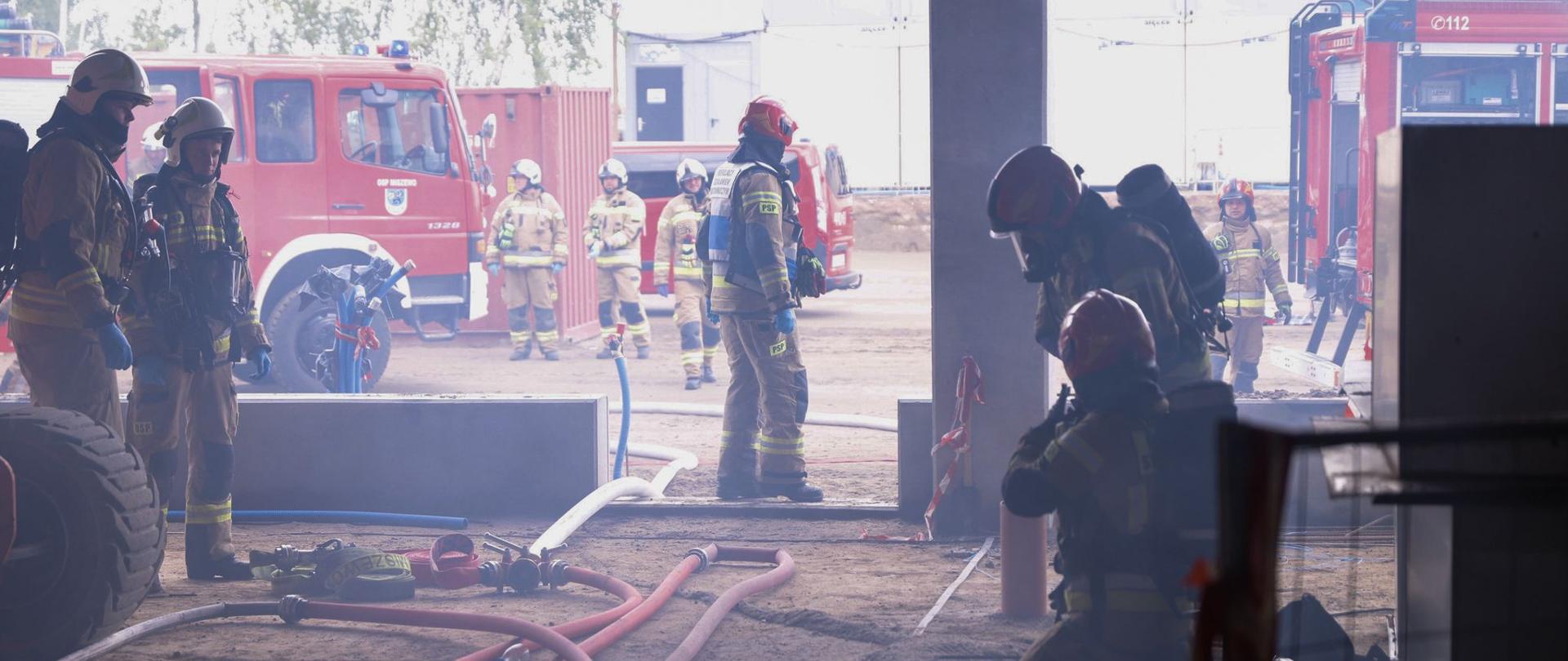 Na zdjęciu widzimy strażaków podczas ćwiczeń - rozłożone linie wężowe, strażacy ubrani w aparaty, w tle samochody. 
