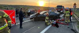 Na pierwszym planie strażak udzielający pomocy poszkodowanej osobie ułożonej na noszach na jezdni przed rozbitym pojazdem. Na drugim planie kolejni strażacy, policjanci, ratownicy medyczni pracujący na miejscu zdarzenia, porozrzucane resztki pojazdów, pojazdy służb.