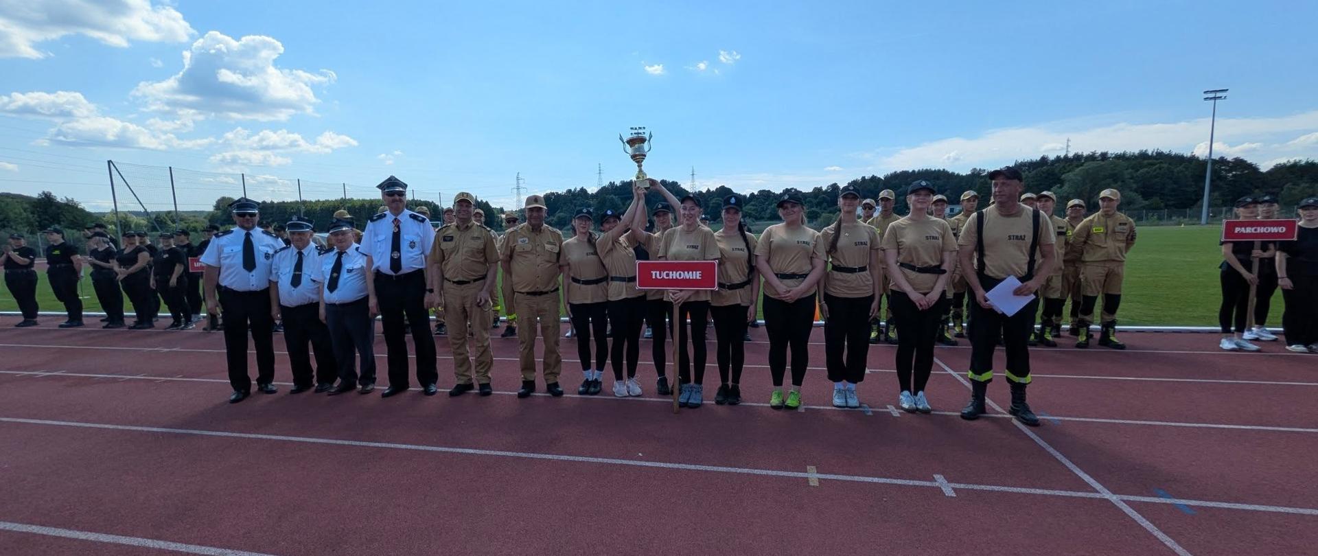 Drużyna kobieca OSP Tuchomie w beżowych koszulkach unosi puchar na podium, otoczona oficjelami i innymi strażakami na bieżni stadionu w słoneczny dzień.