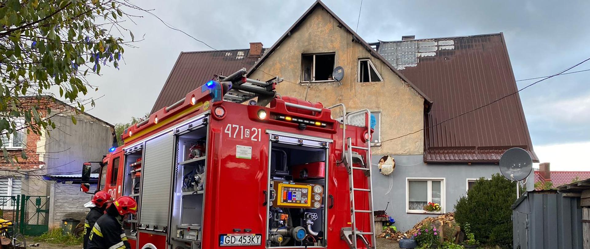 Strażacy przy samochodzie ratowniczo - gaśniczym z rozwiniętymi węzami z samochodu. W tle widok budynku wielorodzinnego po pożarze. Zniszczona drewniana konstrukcja dachu, pokrycie dachowe, 