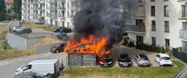 W centralnej części podwórka stoi płonąca wiata śmietnikowa, w tle zabudowa mieszkalna wielorodzinna. Obok wiaty stoi pojazd osobowy, który także zaczyna się palić.