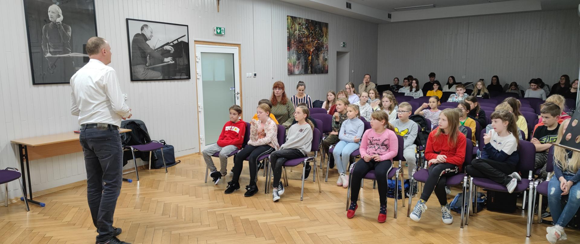 Na zdjęciu widać prowadzącego warsztaty mówiącego do uczniów , który stoi tyłem do obiektywu a przodem do uczniów siedzących na krzesłach na widowni auli szkoły; ściany mają kolor biały a podłoga jest drewniana, jasnobrązowa.