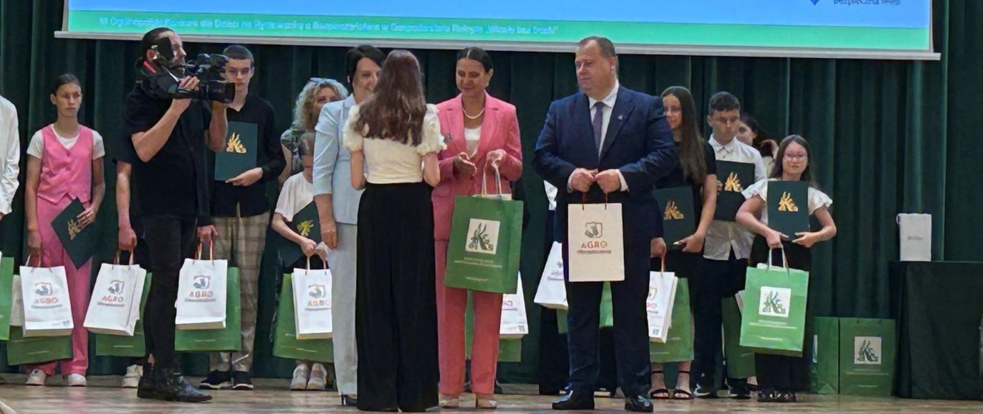 Grupa osób na scenie podczas wręczania nagród. Dwie kobiety w jasnych garniturach wręczają nagrodę nastoletniej dziewczynce. Jedna z kobiet wręcza nastolatce zieloną torbę z logo KRUS.