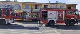 Na zdjęciu widoczne są samochody ratowniczo-gaśnicze, karetka pogotowia oraz budynek mieszkalny wielorodzinny.