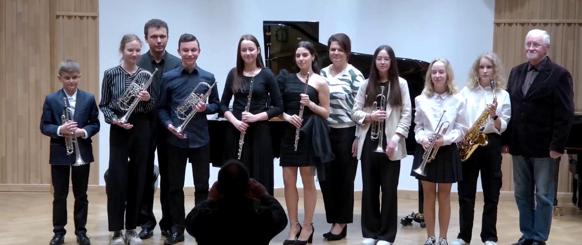 Zdjęcie. Na estradzie stoją wykonawcy koncertu ze swoimi instrumentami dętymi. Pozują do zdjęcia, które wykonuje osoba stojąca przed nimi.