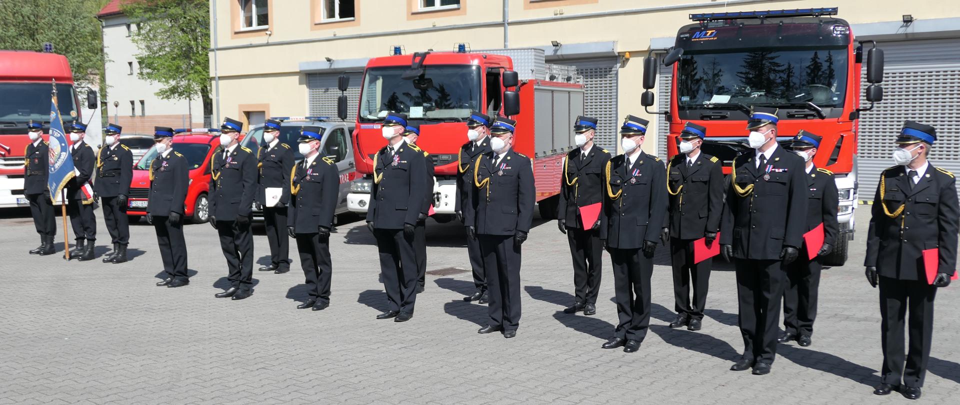 Widok strażaków w ugrupowaniu rozwiniętym w mundurach galowych. W tle samochody pożarnicze i obiekt KP PSP Słubice