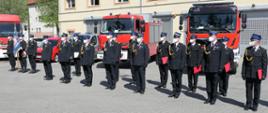 Widok strażaków w ugrupowaniu rozwiniętym w mundurach galowych. W tle samochody pożarnicze i obiekt KP PSP Słubice