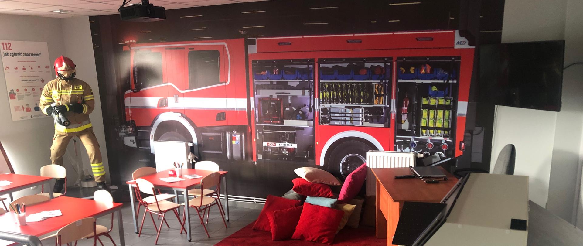 Na zdjęciu na ścianie w 3D samochód ratowniczo gaśniczy na podłodze czerwony dywan z poduszkami stoliki z krzesełkami oraz TV na suficie podłączony rzutnik multimedialny w narożniku manekin strażaka z prądownicą i wężem w ubraniu specjalnym i hełmie 