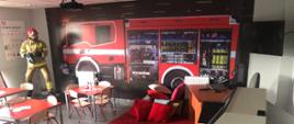 Na zdjęciu na ścianie w 3D samochód ratowniczo gaśniczy na podłodze czerwony dywan z poduszkami stoliki z krzesełkami oraz TV na suficie podłączony rzutnik multimedialny w narożniku manekin strażaka z prądownicą i wężem w ubraniu specjalnym i hełmie 
