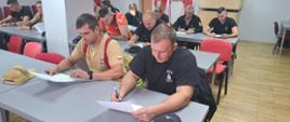 Świetlica komendy powiatowej. Przy stołach siedzą uczestnicy szkolenia, którzy rozwiązują test pisemny. W tle widoczny jest stojak z flagami biało-czerwonymi oraz ścianka promocyjna.