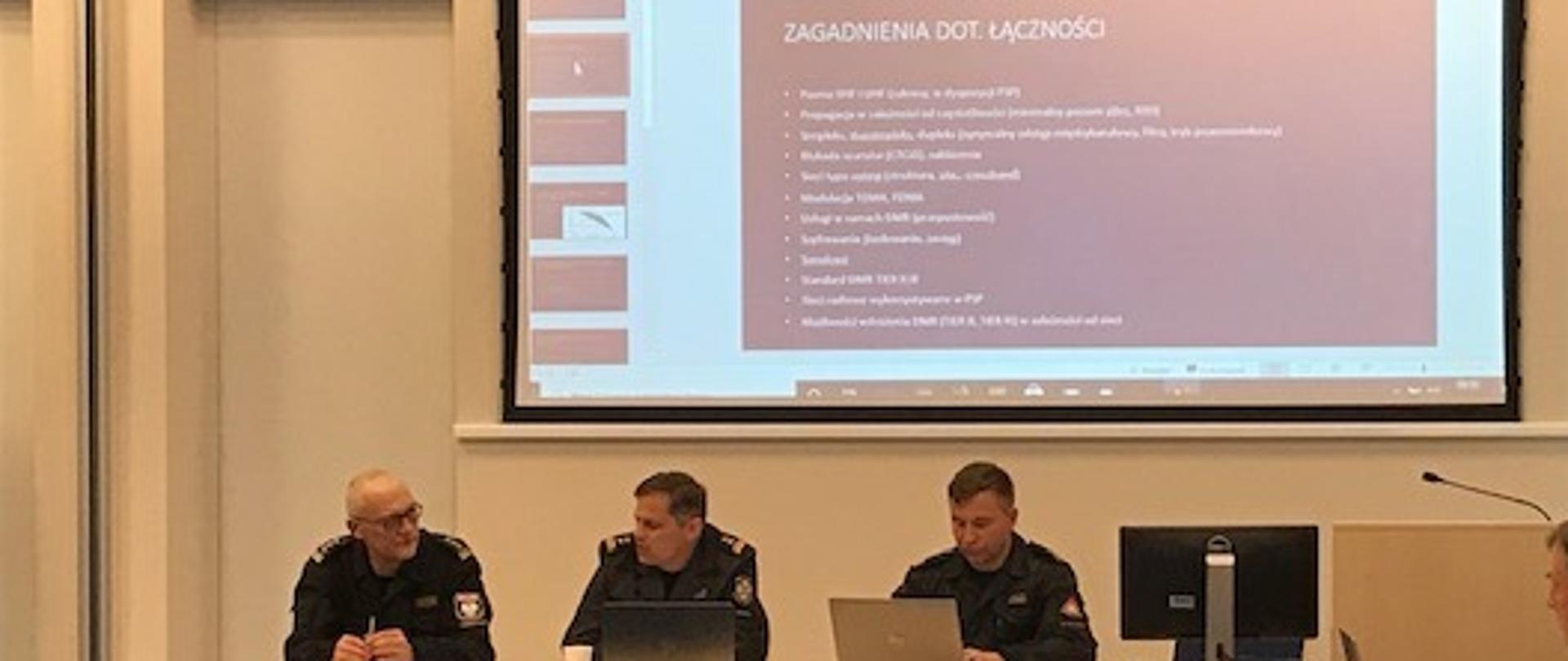 Trzech funkcjonariuszy PSP siedzi przy stole prowadząc spotkanie , w tle slajd z prezentacji