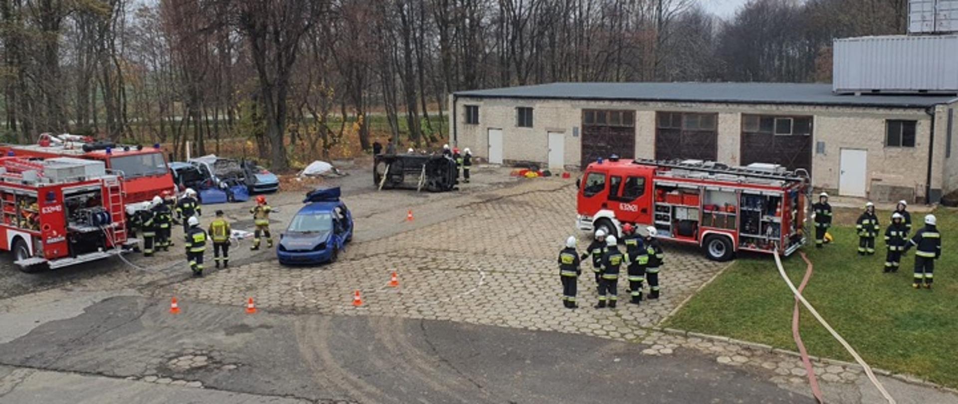Strażacy z OSP podczas egzaminu praktycznego. Z lewej strony prowadzone są działania ratownicze polegające na wykonaniu dostępu do osoby poszkodowanej w samochodzie osobowym. Po prawej stronie budowanie linii gaśniczych. 