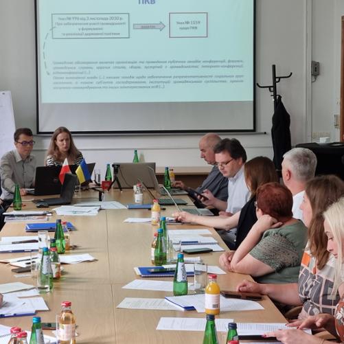 W sali przy stole siedzą uczestnicy i patrzą się na materiały szkoleniowe leżące na stole i wyświetlane na ekranie