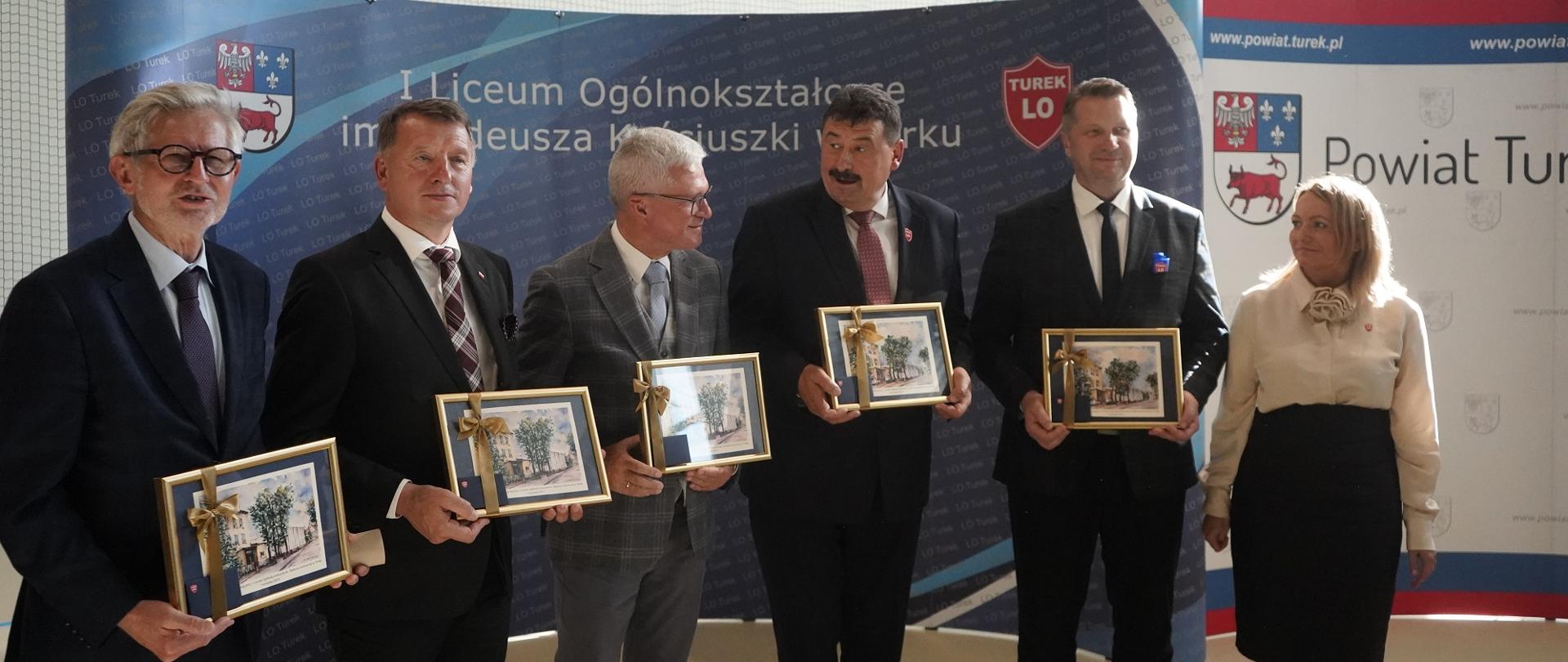 Minister Czarnek, czterech mężczyzn w garniturach i kobieta w białej bluzce stoją w szeregu i trzymają w rękach oprawione w ramki dokumenty.