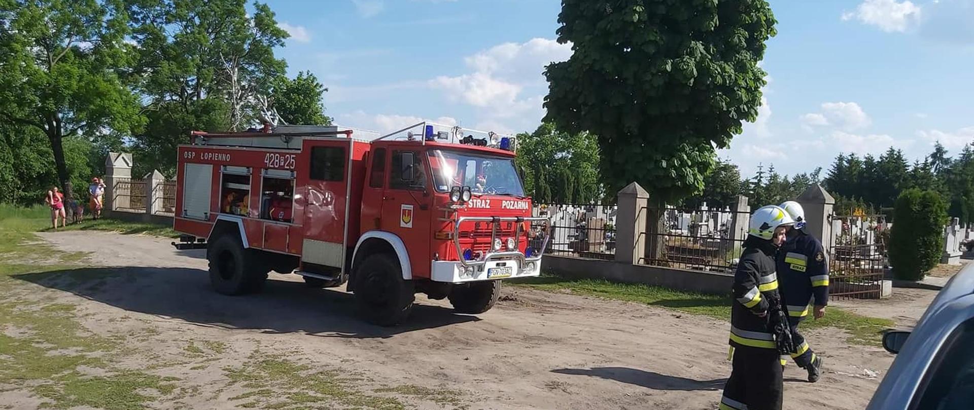 Słoneczny dzień. Czerwony wóz strażacki stoi przed bramą cmentarza na piaszczystej drodze. W tle szare nagrobki i zielone wysokie drzewa. Dwóch strażaków w czarnych ubraniach i białych hełmach stoi przed otwartą bramą.