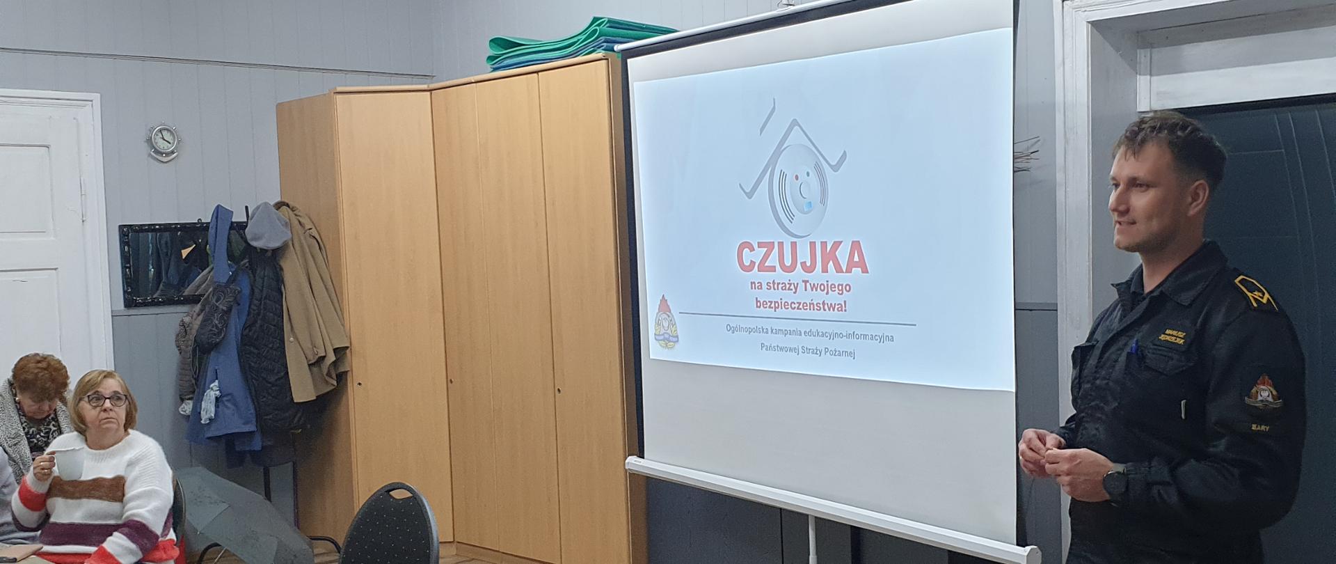 Strażak przeprowadza pogadankę z seniorami na temat bezpieczeństwa zakresie ochrony przeciwpożarowej, czujek i pożarów.