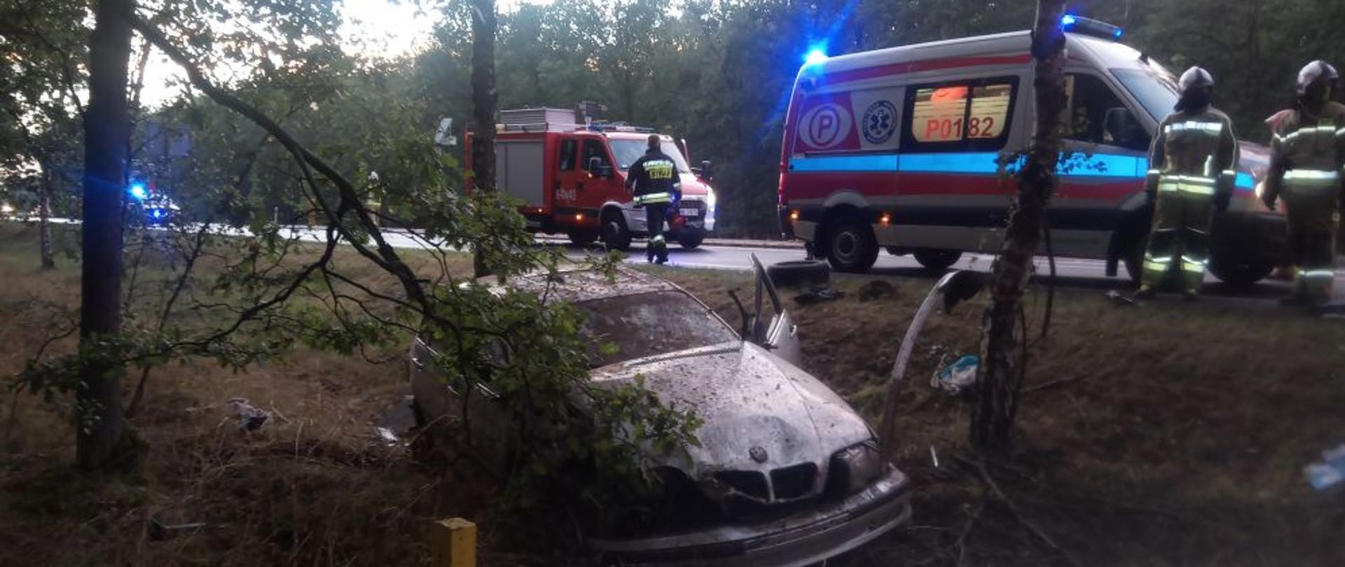 Rozbity samochód osobowy znajdujący się w rowie. W tle, Zespół Ratownictwa Medycznego oraz zastęp strażaków. Sytuacji przyglądają się druhowie z OSP Kębłowo.