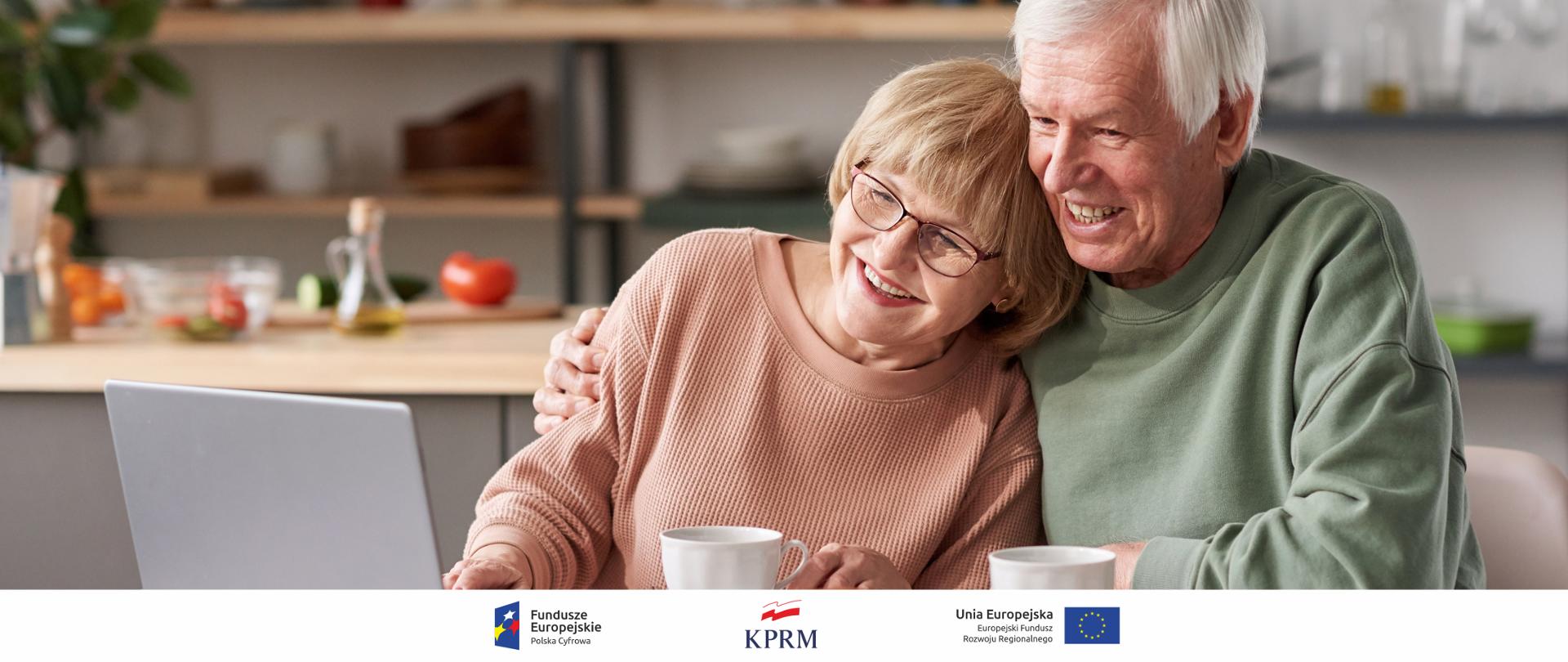 Dwoje starszych osób siedzą objęci i uśmiechnięci za kuchennym stołem, patrzą w ekran laptopa.