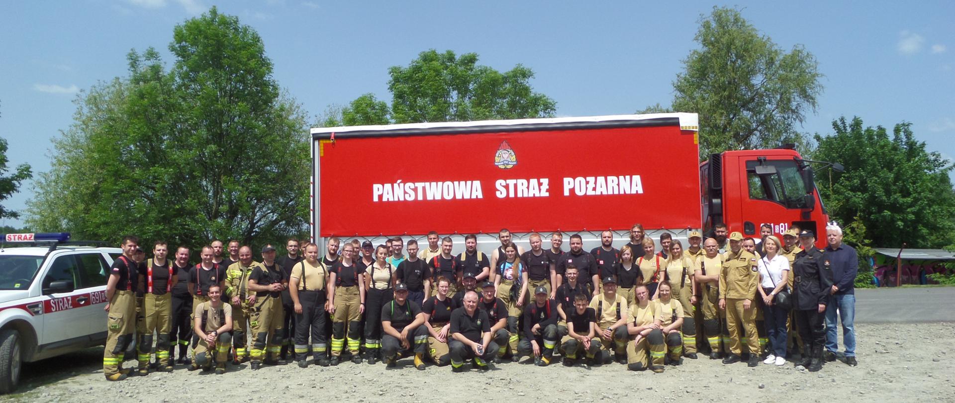 Zdjęcie grupowe wszystkich uczestników ćwiczeń. W tle samochód strażacki z napisem na czerwonej plandece Państwowa Straż Pożarna i logo PSP. Za nim drzewa.