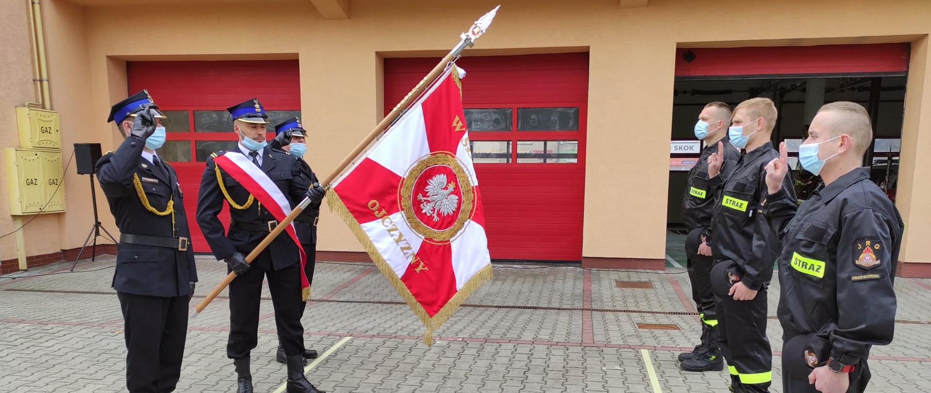Fotografia przedstawia ślubowanie młodych mężczyzn przyjętych do służby.
Po lewej stronie jest poczet sztandarowy, składający się z trzech mężczyzn w strojach wyjściowych a na ich ramionach przewieszony jest szósty sznur.
Dwoje oddaje honor poprzez wyprostowane dwa palce zbliżone do brzegu daszka czapki.
Środkowy trzyma w rękach pochylony sztandar a jego lewa noga jest wysunięta do przodu.
Po prawej stronie fotografii stoją młodzi strażacy w trakcie ślubowania.
Ubrani są w czarne stroje straży pożarnej, prawą rękę mają zgiętą do ślubowania zaś w wyprostowanej lewej trzymają czapki.