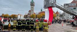 Strażacy Państwowej Straży Pożarnej oddają honor podczas podnoszenia flagi państwowej, obok stoją mieszkańcy miasta.