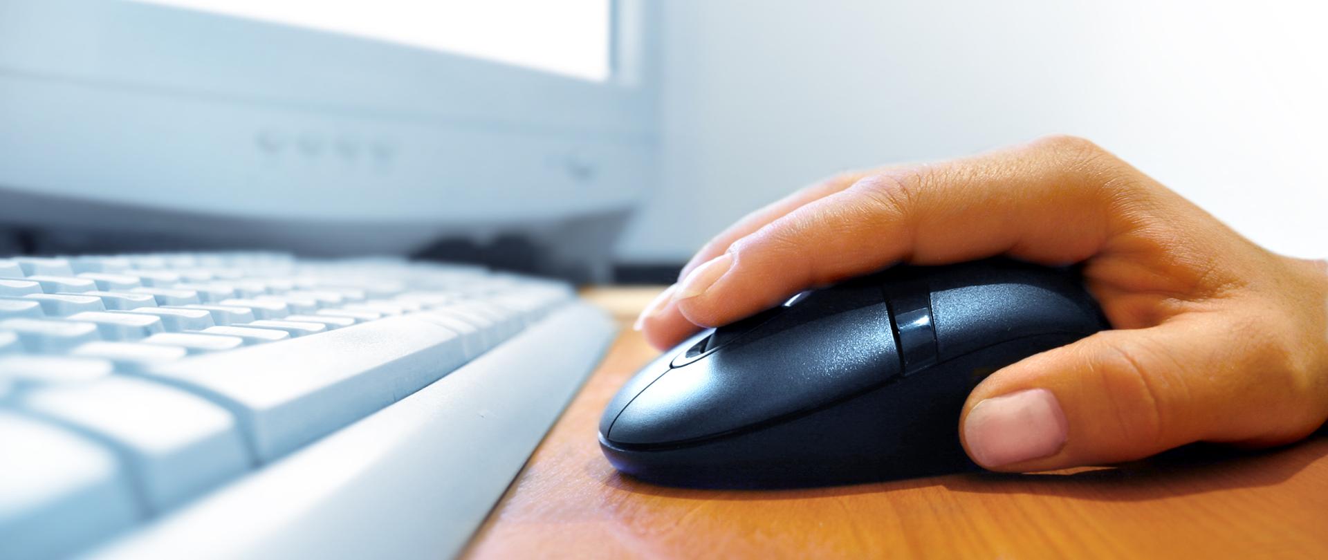 Na obrazku widać dłoń trzymającą mysz komputerową, przed nią klawiatura i monitor.