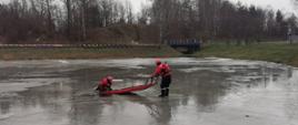 Fotografia przedstawiająca ćwiczenia strażaków na lodzie gdzie strażak wyciąga poszkodowanego z wody za pomocą sań lodowych.