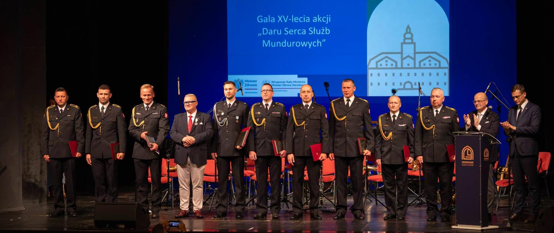 Strażacy w mundurach galowych stojący w szeregu na scenie podczas uroczystej gali