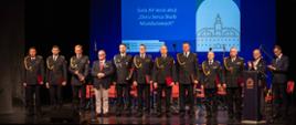 Strażacy w mundurach galowych stojący w szeregu na scenie podczas uroczystej gali