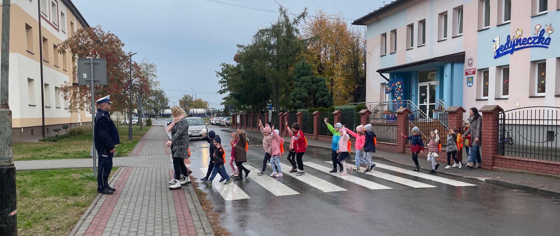 Dzieci przechodzą przez jezdnię na pasach. Mają podniesione ręce. Pada deszcz. Widać samochód. 