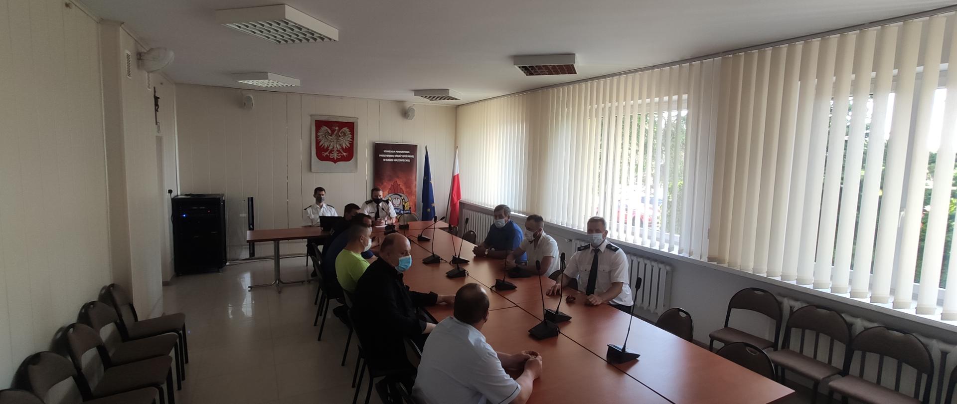 Zdjęcie przedstawia 9 mężczyzn siedzących za stołem, w kształcie litery T. Za dwójką z nich na ścianie znajduje się godło polski oraz roll-up z logo straży pożarnej. Ci dwaj mężczyźni ubrani są w mundur strażacki, reszta mężczyzn, siedząca przy stołach prostopadle do tej dwójki, ubrana jest po cywilu. Na stole znajdują się mikrofony.