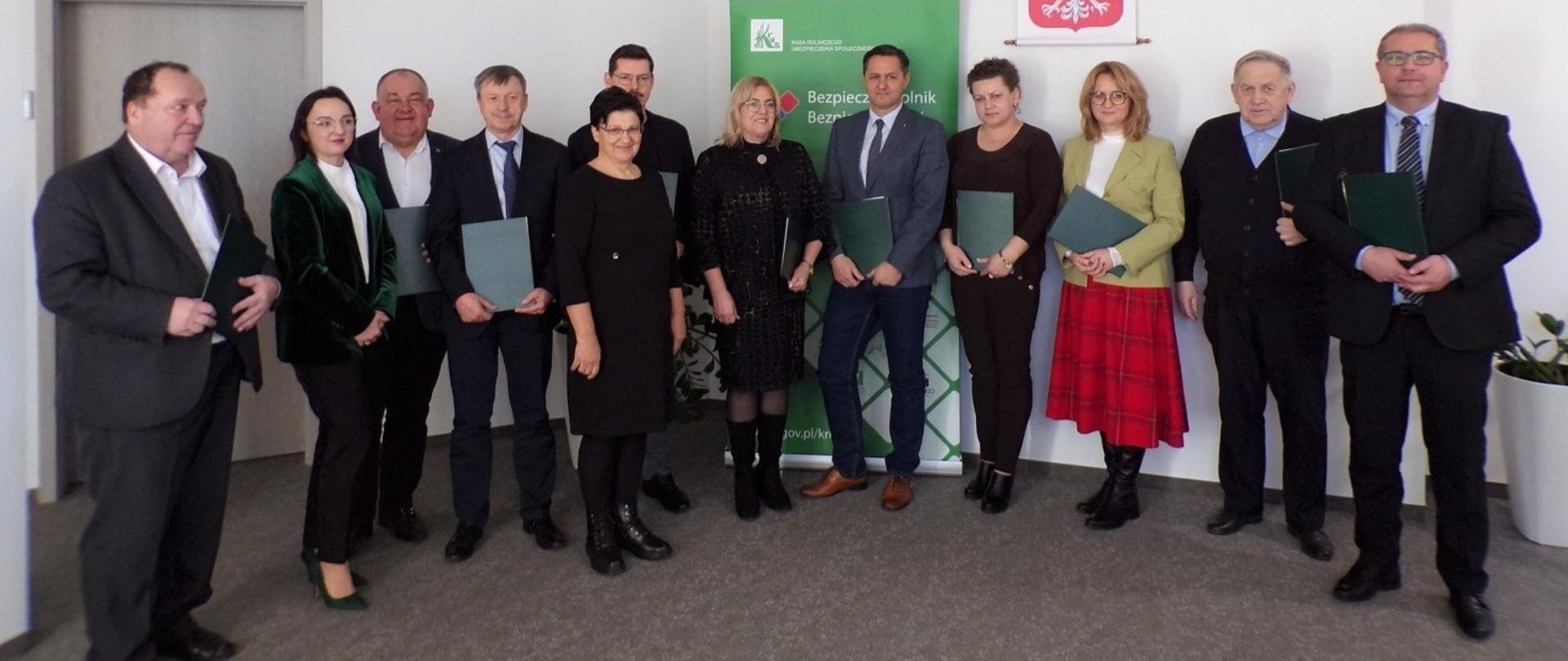 Pięć kobiet i siedmiu mężczyzn pozuje do zdjęcia na tle roll-upu KRUS i godła. Wszyscy trzymają w dłoniach zielone teczki z tekstem porozumienia i uśmiechają się do zdjęcia.