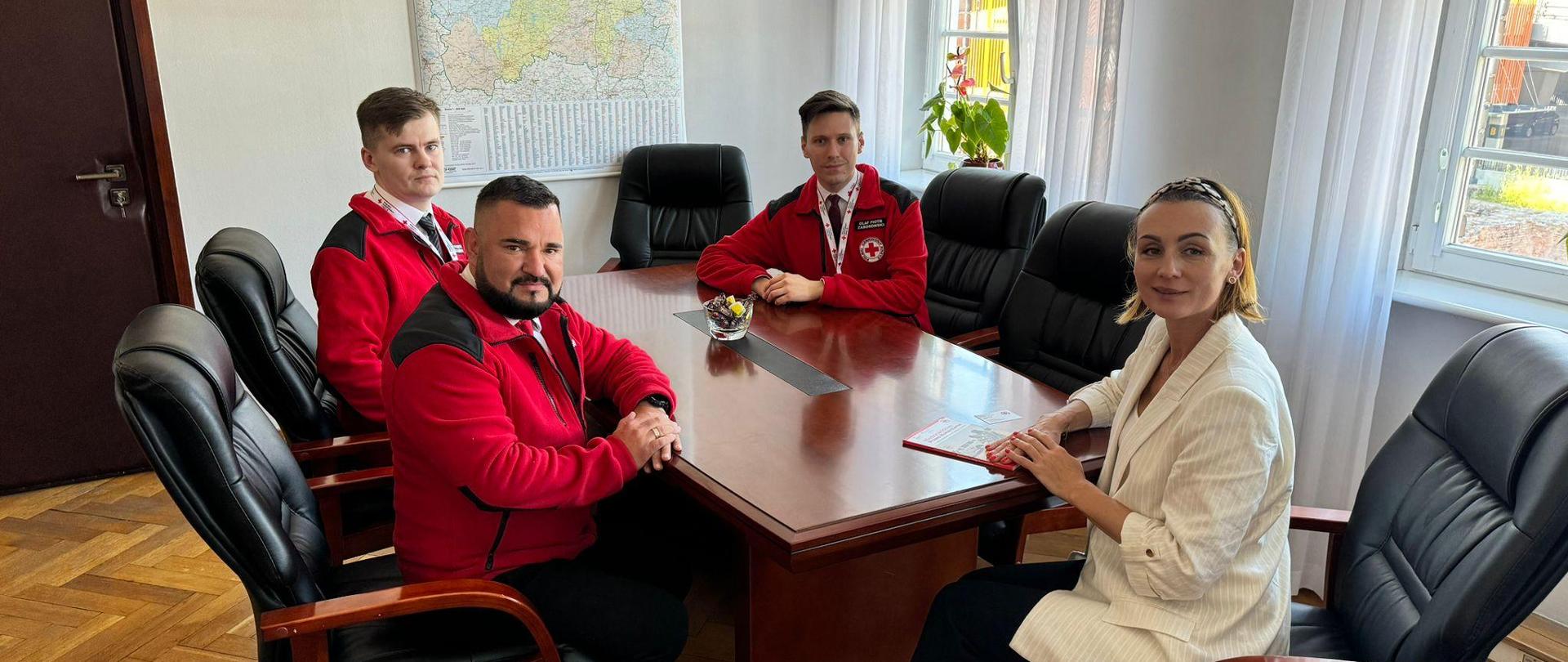 Grupa osób - kobieta (wojewoda) i kilku mężczyzn w strojach ratowników siedzą przy stole i rozmawiają