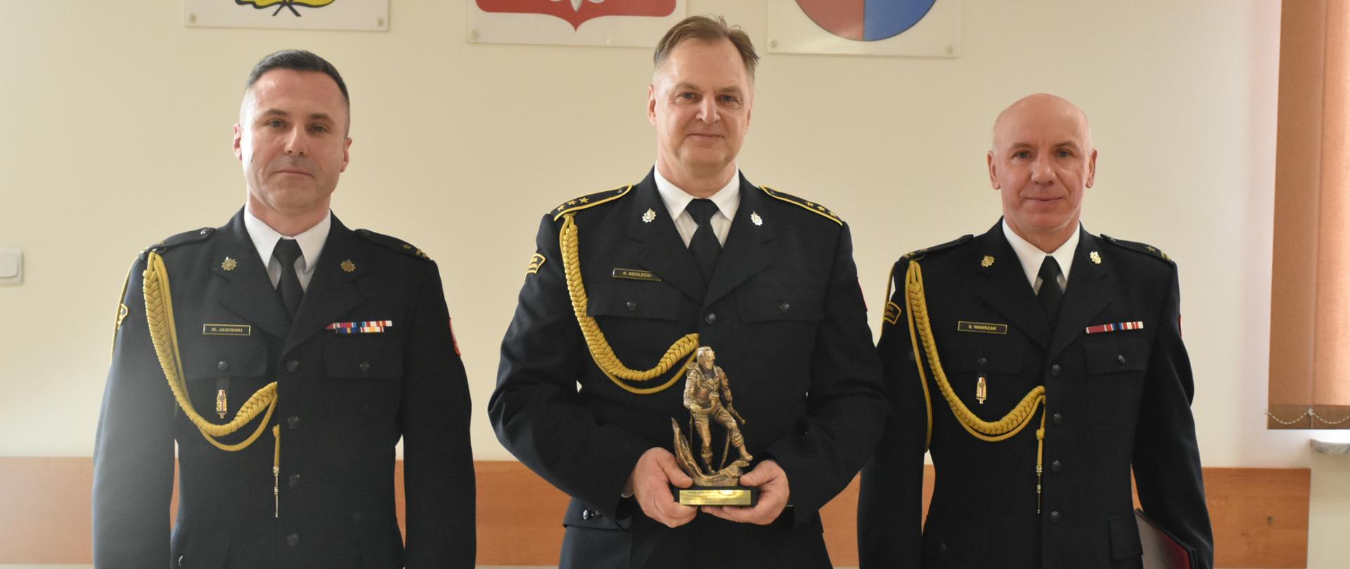 Zdjęcie komendanta oraz zastępcy z odchodzącym na emeryturę z-cą dowódcy jednostki. W tle widać Godło Polski, herb powiatu oraz PSP.