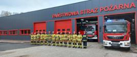 Przed garażem Jednostki Ratowniczo-Gaśniczej strażacy ubrani w ubrania specjalne stojąc w szeregu oddają hołd, w tle samochody ratowniczo - gaśnicze