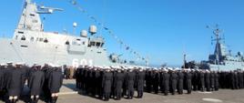 Żołnierze Marynarki Wojennej RP stoją na nabrzeżu w rzędach przed nimi zacumowany jest okręt wojenny.