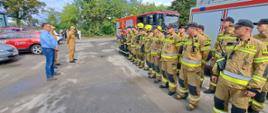 Na zdjęciu widzimy strażaków, podczas odprawy po ćwiczeniach stojących w dwuszeregu na tle samochodów gaśniczych.