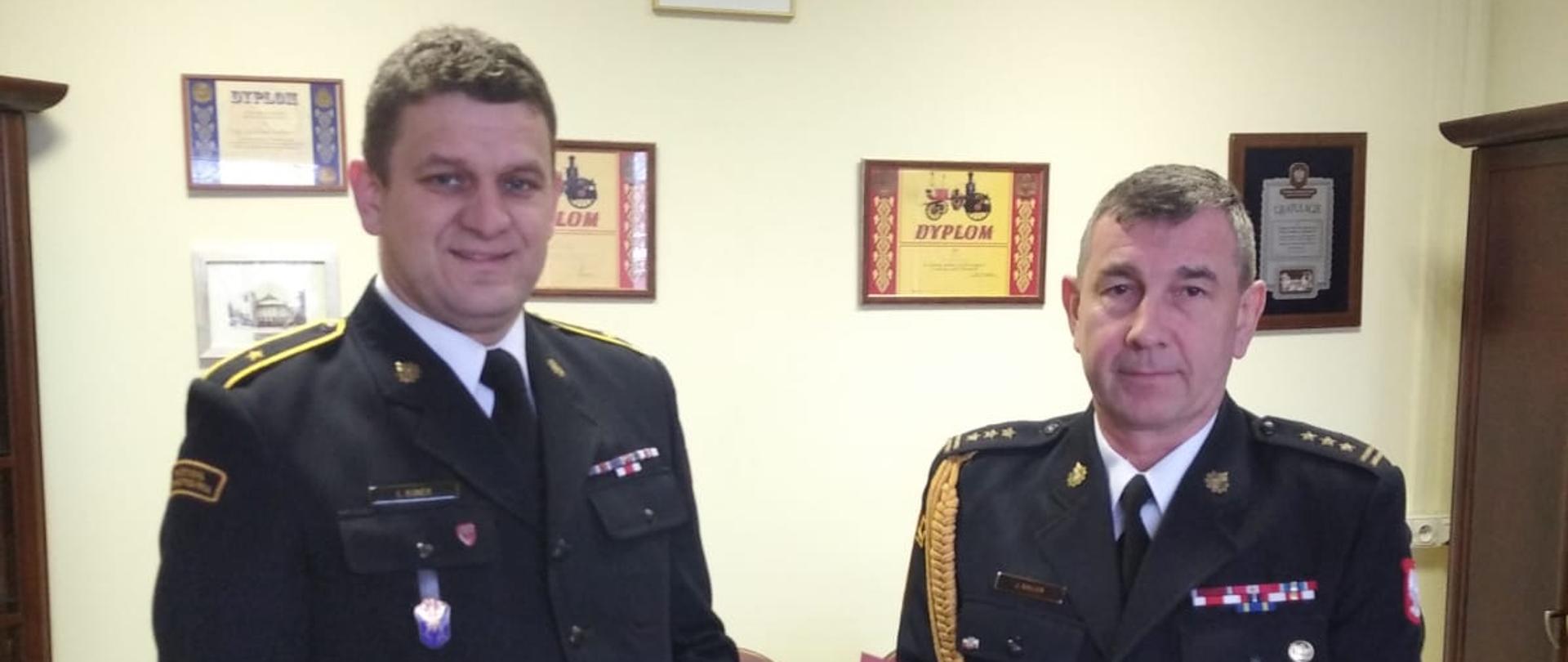 Z lewej asp. Łukasz Kunek trzymający książkę z zieloną okładką, z prawej st. bryg. Janisław Buller trzymający teczkę z dyplomem. Obydwaj w mundurach granatowych wyjściowych znajdują się w gabinecie Komendanta, w tle żółta ściana z dyplomami i godłem RP.