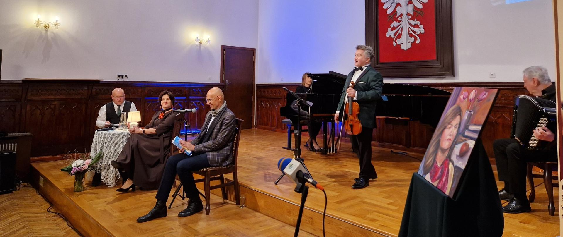 Na scenie Sali Królewskiej odbywa się benefis poetki Aleksandry Piguły. Troje prowadzących siedzi przy stole z lampką, a obok występują muzycy – śpiewak, pianistka i akordeonista. W tle widoczne są portrety i duży herb z białym orłem.