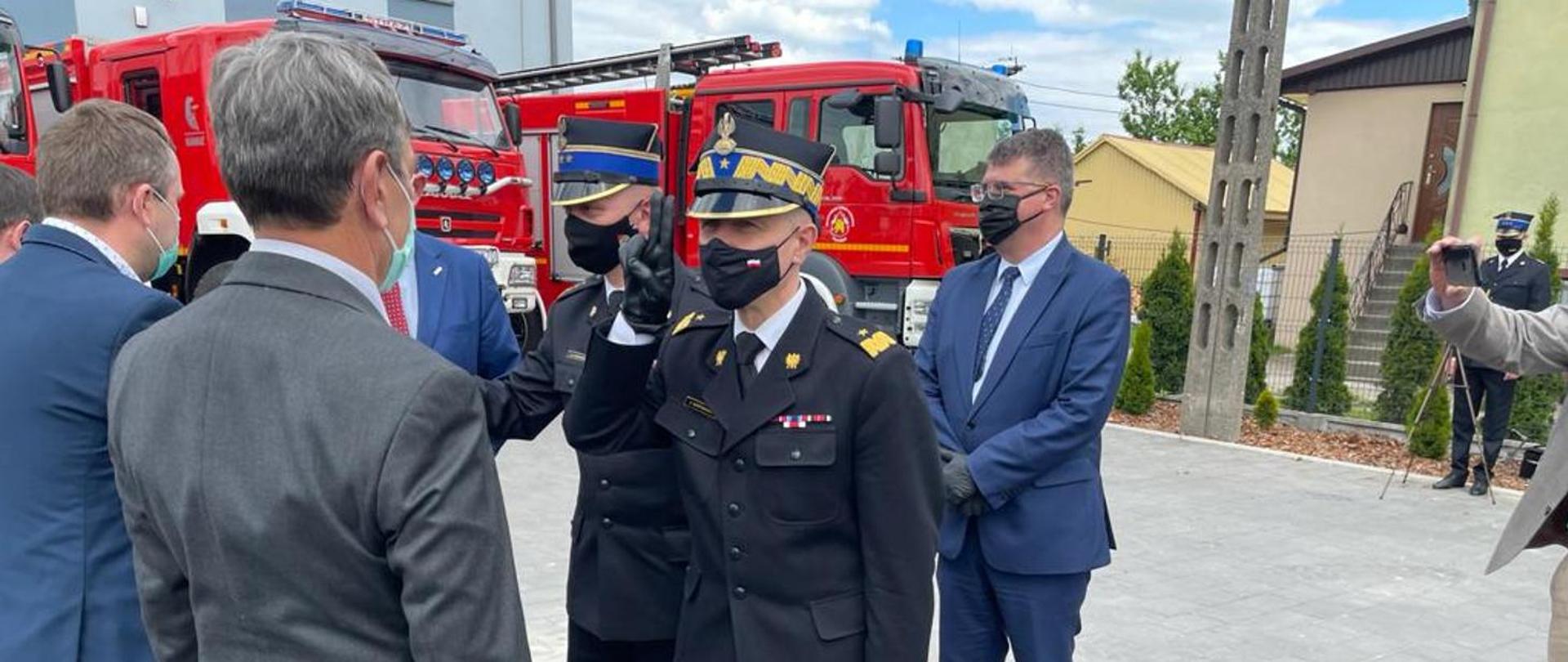 Zdjęcie przedstawia przekazanie promes przedstawicielom jednostek OSP. Komendant Główny Państwowej Straży Pożarnej w umundurowaniu galowym, oddaje honor w trakcie wręczania promes. W tle Wozy strażackie oraz Wiceminister.